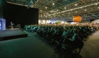 Large seated crowd within an exhibition hall listening to an engaging talk.