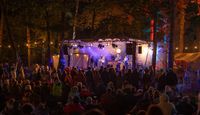 Evening photo of Between the Trees Festival in Wales, showing small stage in the woods with musicians playing to an appreciative crowd.