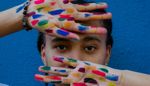 Young person with the palms of their hands facing out in front of their face, the palms are painted in colourful dashes.