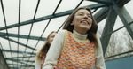Film still showing a smiling woman beneath the glass and steel roof of a covered walkway.