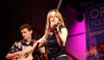 Folk musician performing on stage with violin, with guitarist in the background.