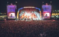 A large crowd at night gathered for Boardmasters outdoor music festival, with the stage lit up by vibrant lights and pyrotechnics, flanked by two large screens displaying the event's name, Boardmasters, and sponsor logos.