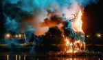 Large burning stag sculpture surrounded by coloured smoke at night during Lost Village Festival, reflected in the water below.