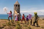 Performers in colourful costumes walk in a line across a coastal hillside near a stone tower at Ventnor Fringe, with the sea and cliffs visible in the background.