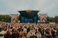 Crowd at Parklife Festival facing the main stage with large screens showing cheering festival-goers, set in Heaton Park with trees in the background.