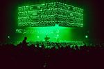 Crowd facing a green-lit electronic music stage with large-scale video screens.