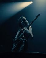 Guitarist performing alone beneath a white spotlight on a dark indoor stage.