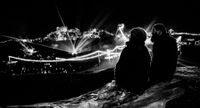 Two people silhouetted at night overlooking Equinox Festival site illuminated by string lights and laser beams, with a scenic backdrop of trees and structures.