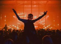 Terminal V Halloween Festival goer with luminous painted face where the eyes are x's and mouth simple lines, surrounded by vibrant lights on the stage in the background.