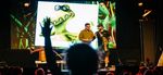 Two presenters on stage in front of an illustrated dinosaur while audience members raise their hands