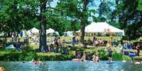 ALSO Festival, a vibrant summer festival scene by a lake with people swimming, floating on inflatables, and relaxing on the grass under the shade of large trees. A marquee with a 'BAR' sign stands prominently in the background amidst the lush greenery on a sunny day.