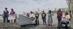 A small white boat sits on the shore just beyond the waves, a multinational group of women stand in a semi circle around a small white boat.