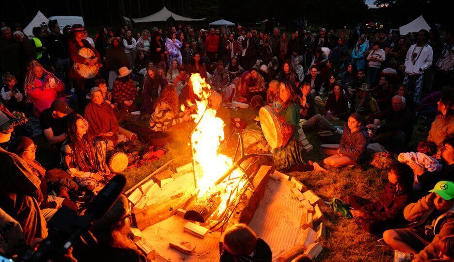 Gathering around the Soul Revolution Festival fire, goers playing drums and adding wood to fire, festival crowd surrounding the space.