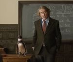 Film still showing a suited man standing beside a penguin in front of a classroom blackboard.