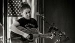 Black-and-white photograph of a woman playing acoustic guitar beside a microphone, with an American flag behind her. Photograph: Tim Mossholder via Unsplash.