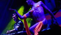 Performer dancing on stage in vibrant orange costume beside a DJ at Mighty Hoopla, illuminated by colourful festival lighting.