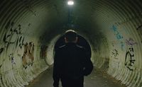 A film still image of person walking down a graffitied corrugated tunnel.