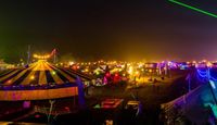 Two people silhouetted at night overlooking Equinox Festival site illuminated by string lights and laser beams, with a scenic backdrop of trees and structures.