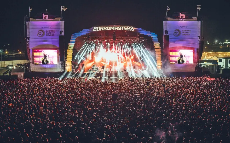 A large crowd at night gathered for Boardmasters outdoor music festival, with the stage lit up by vibrant lights and pyrotechnics, flanked by two large screens displaying the event's name, Boardmasters, and sponsor logos.