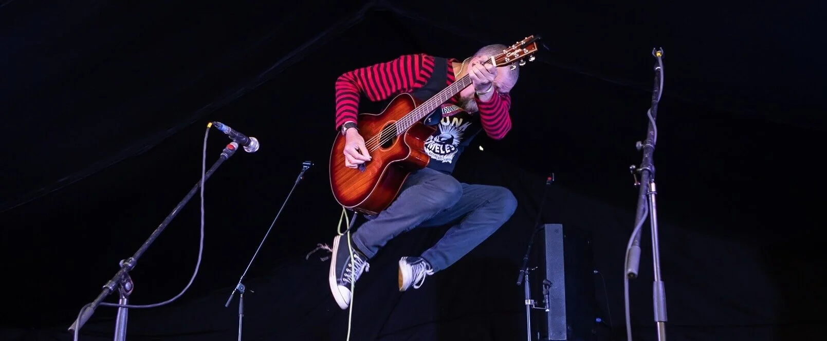 A dynamic performer in a striped red and black sweater and blue pants playing an acoustic guitar mid-jump on a dimly lit stage, with microphones and stands in the foreground against a dark background.