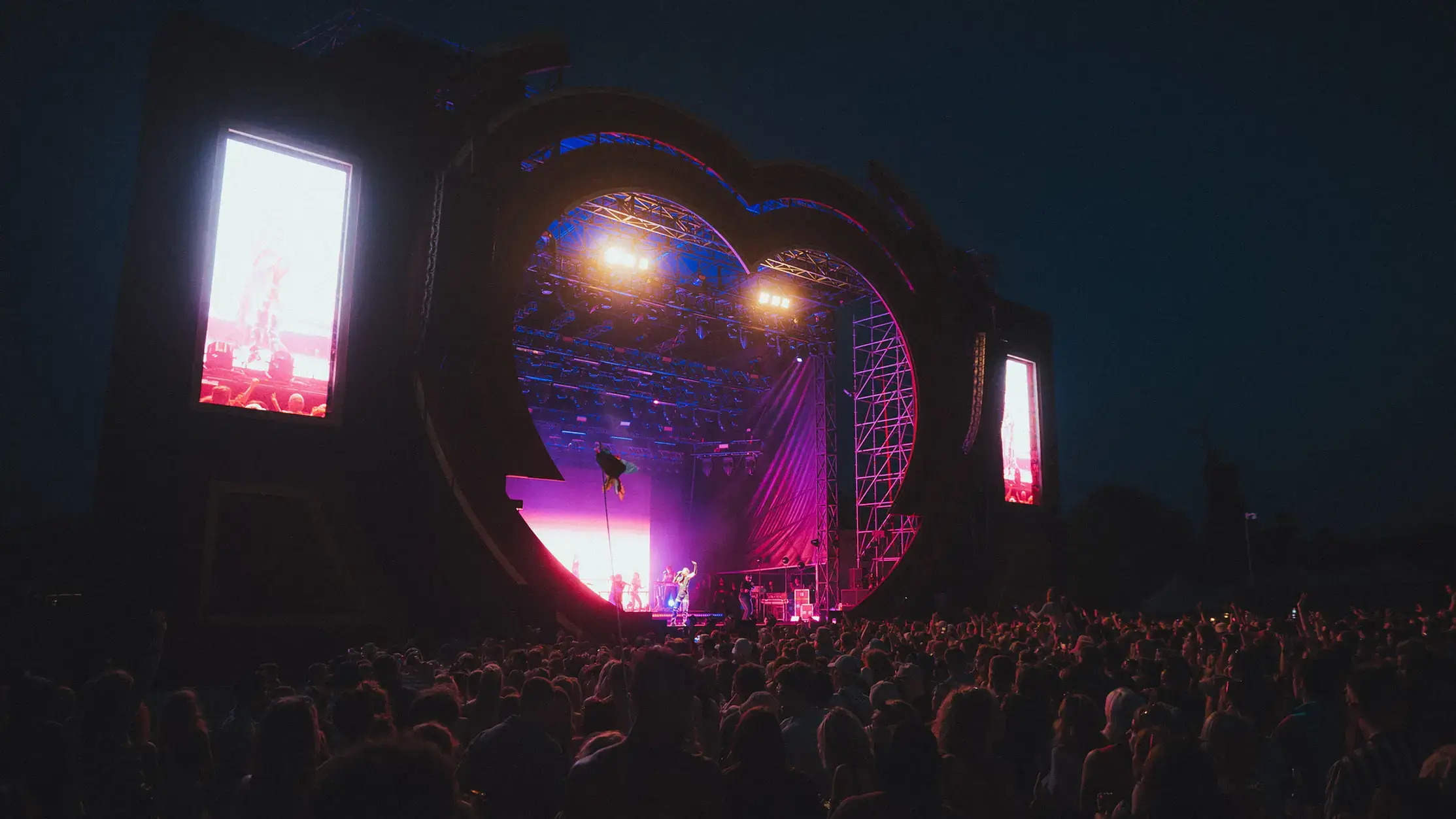 A large crowd gathered at Love Saves The Day music festival during twilight. The stage is set with a unique heart-shaped arch, lit with vibrant stage lights and flanked by large screens displaying performers. The atmosphere is lively with festival-goers enjoying the live music.