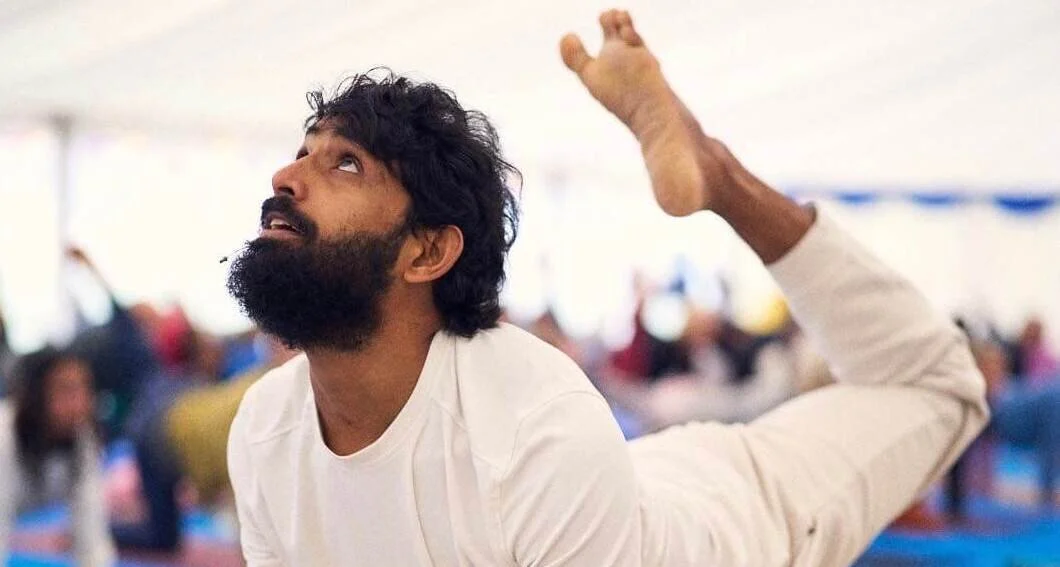 A man with a beard practicing a yoga pose indoors, raising one leg up with a focused expression, surrounded by other participants in the background.